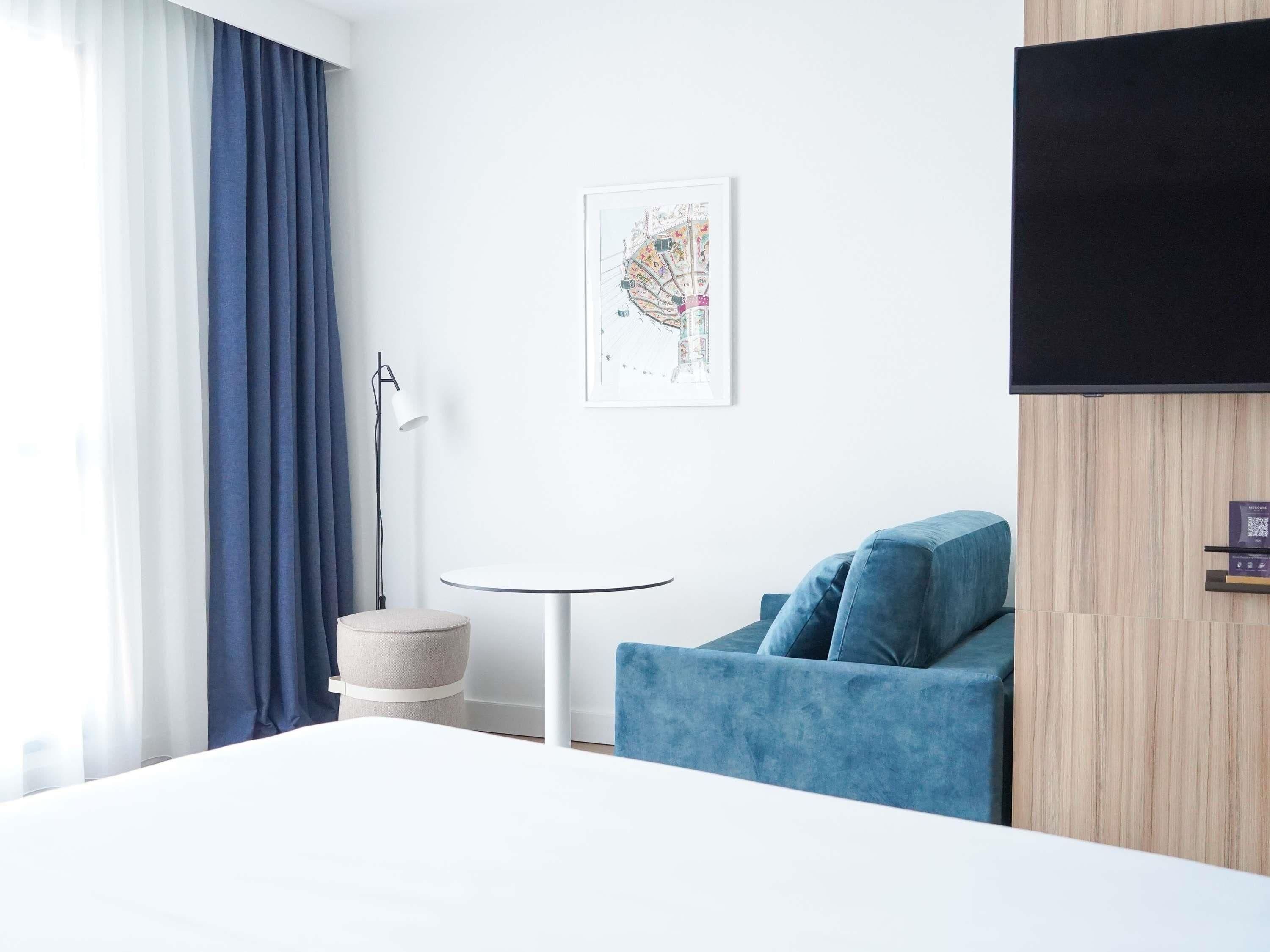 A modern hotel room with a bed, a blue armchair, a small round table, a lamp, a wall-mounted TV, and a framed map.