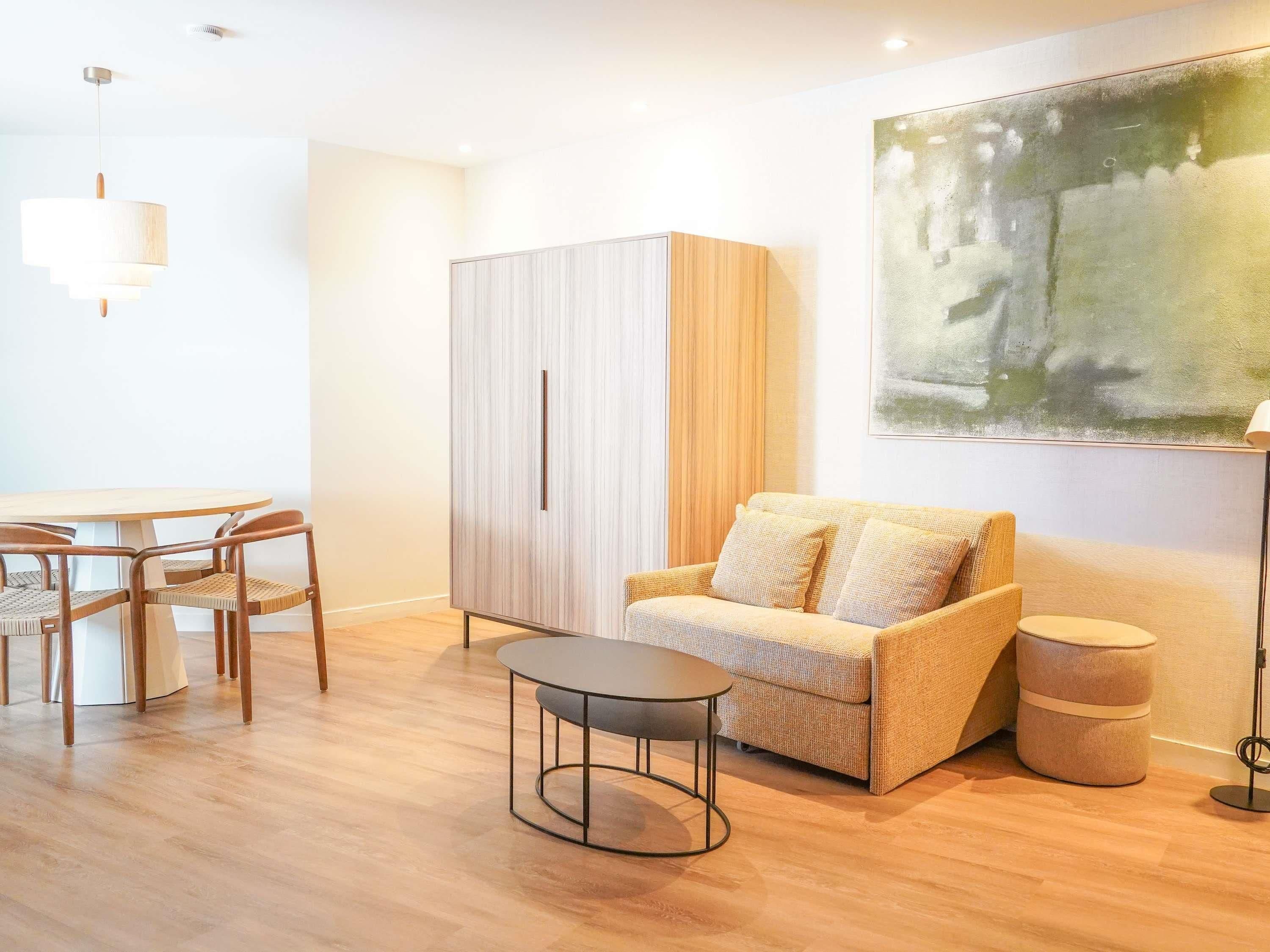 A modern living room with a wooden floor, a round table with chairs, a sofa, a cabinet, and a painting on the wall.