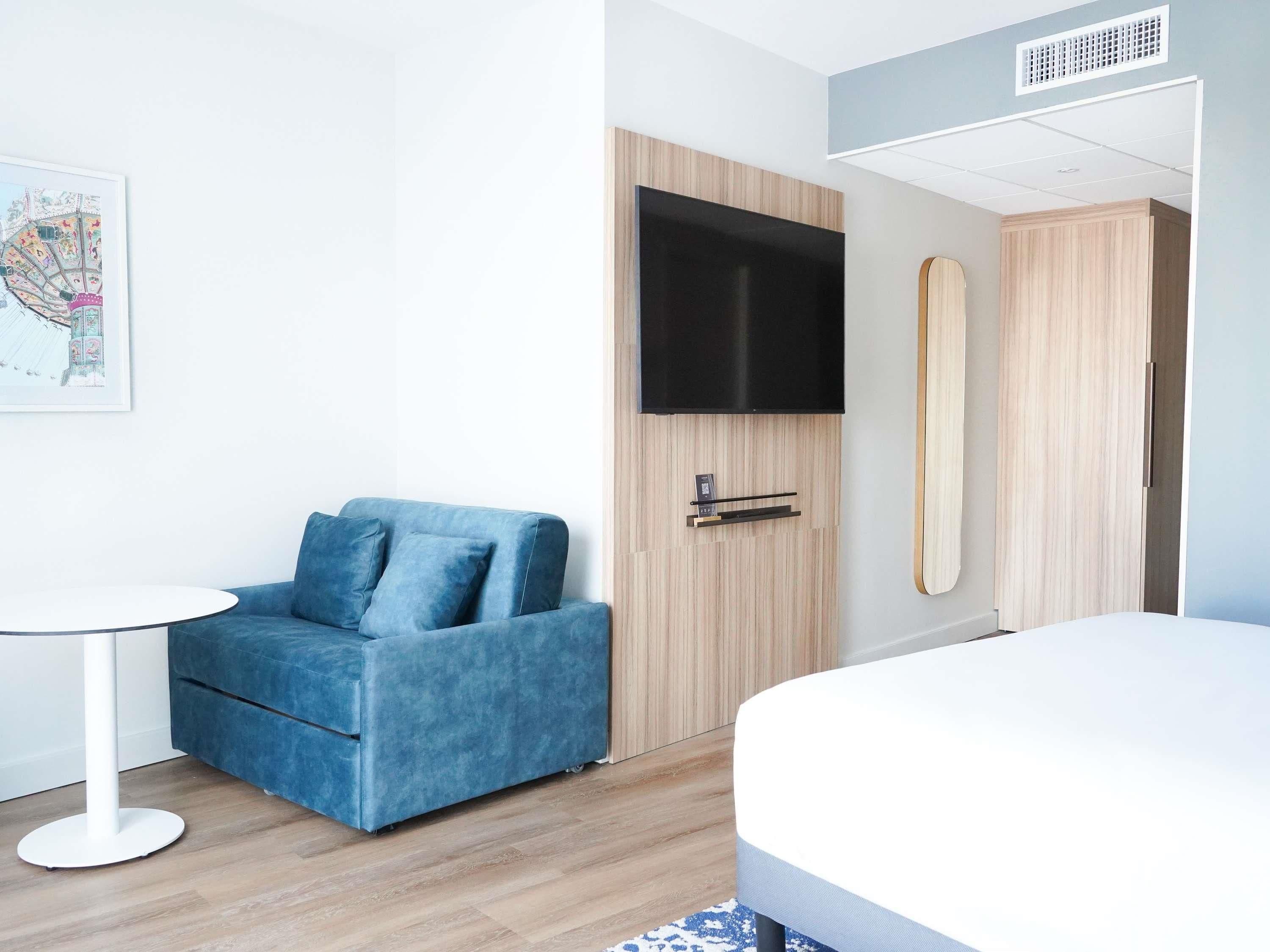 Ein modernes Hotelzimmer mit blauem Sofa, einem Bett, einem Flachbildfernseher und einem Holzkleiderschrank.
