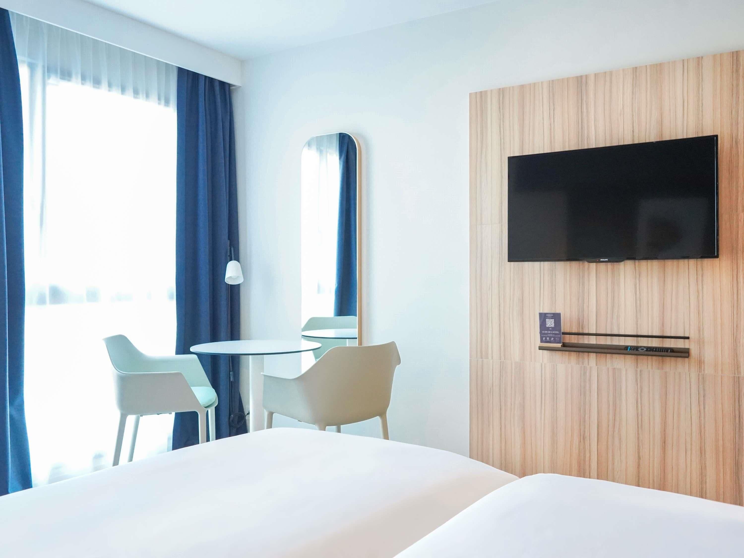 Habitación de hotel con cama, televisión montada en la pared, una pequeña mesa con sillas y una ventana con cortinas.