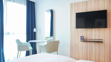 Une chambre d’hôtel avec un lit, un téléviseur fixé au mur, une petite table avec des chaises et une fenêtre avec des rideaux.