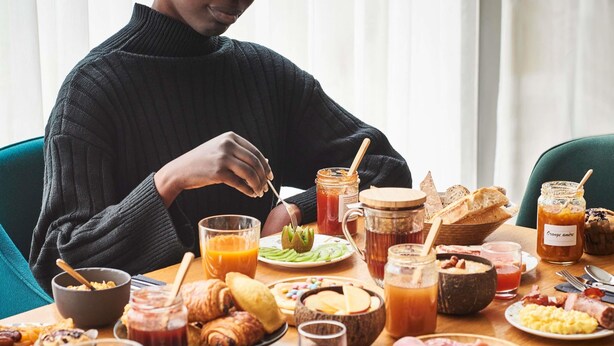 A breakfast table with various food items, a person eating, and a window in the background.
