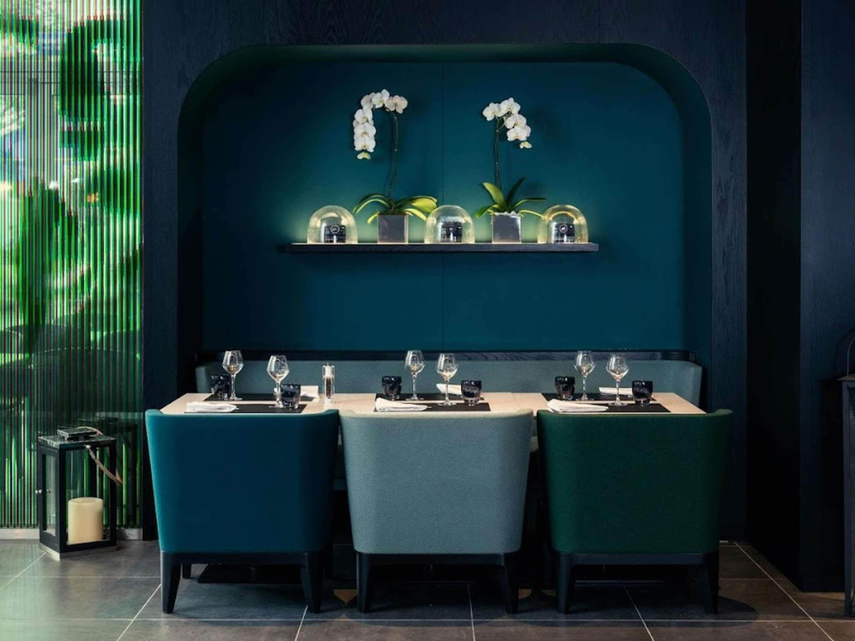 A restaurant with a teal-colored wall, a row of tables with chairs, and a display of orchids and vases on the wall.