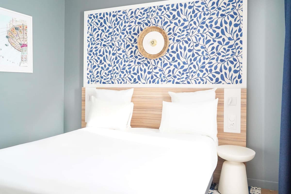 A bedroom with a bed, white bedding, a wooden headboard, a wall-mounted clock, and a small round table.