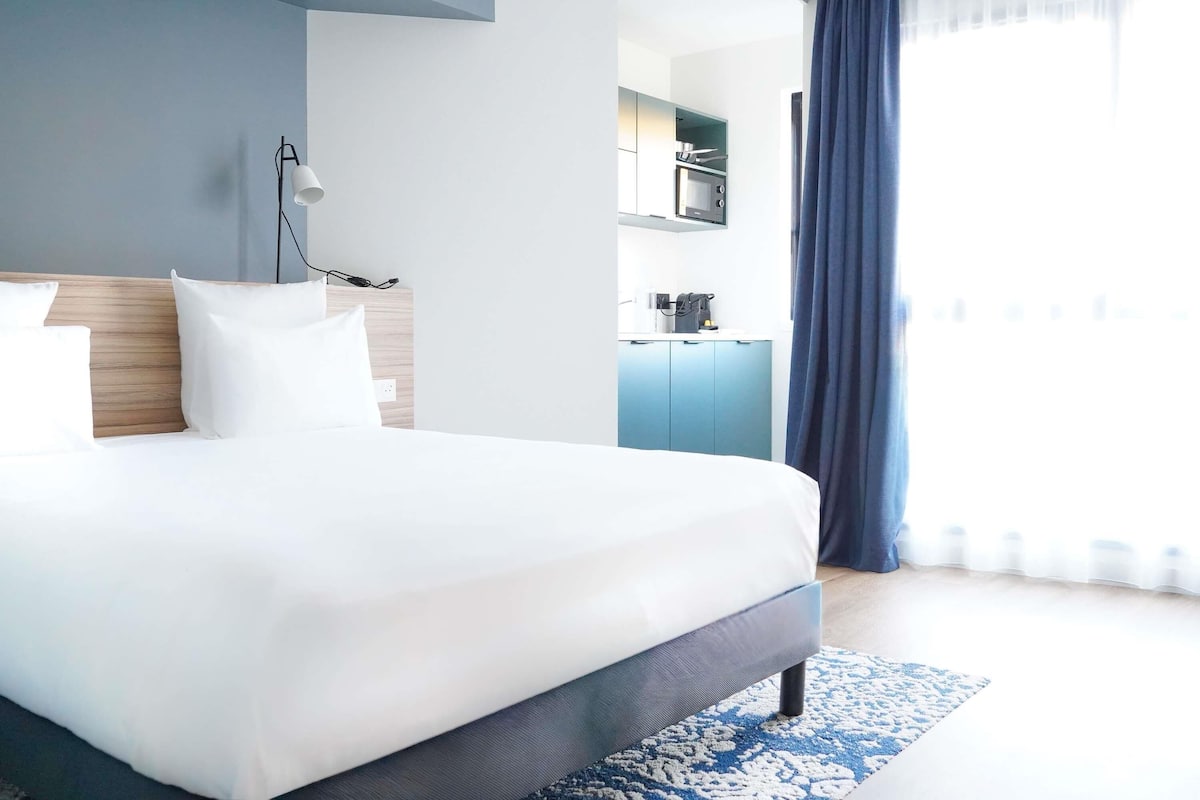 A modern hotel room with a large bed, a desk with a computer, a kitchenette, and a view of the city.