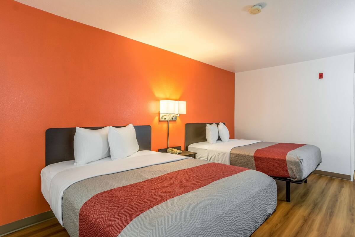 A hotel room with two beds, a nightstand, a lamp, and a wooden floor.