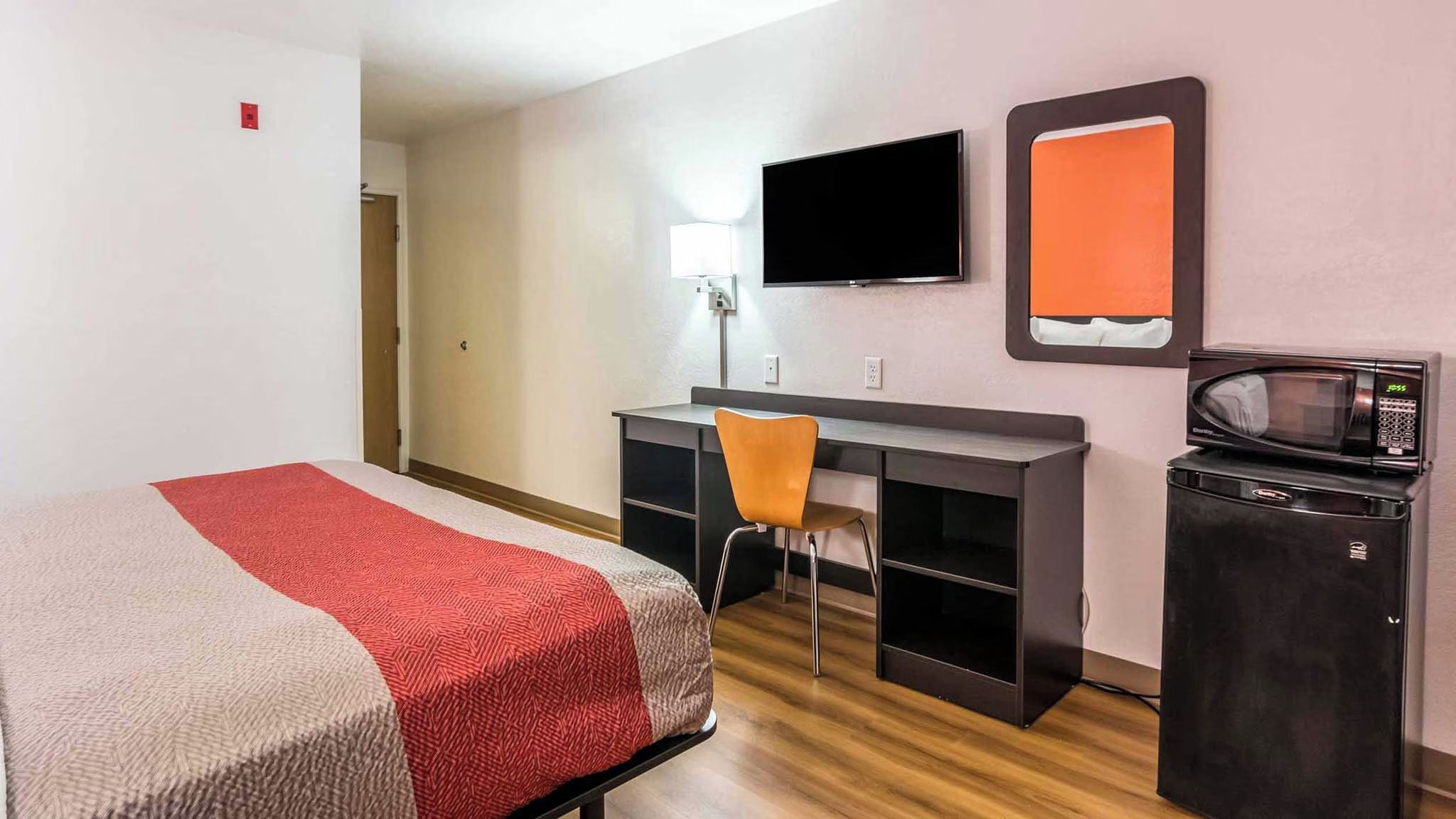 Quarto de hotel com cama, escrivaninha, cadeira, televisão, micro-ondas e pequeno frigorífico.