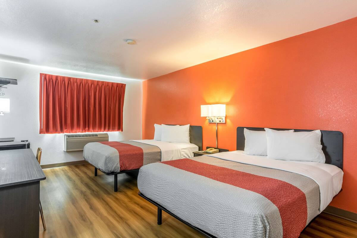 A hotel room with two beds, a desk, and a window with red curtains.