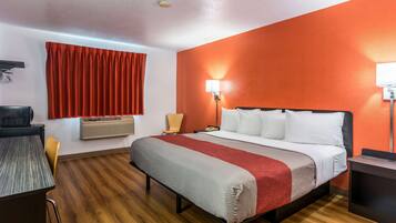 A hotel room with a bed, a desk, a chair, a window with red curtains, and an air conditioning unit.