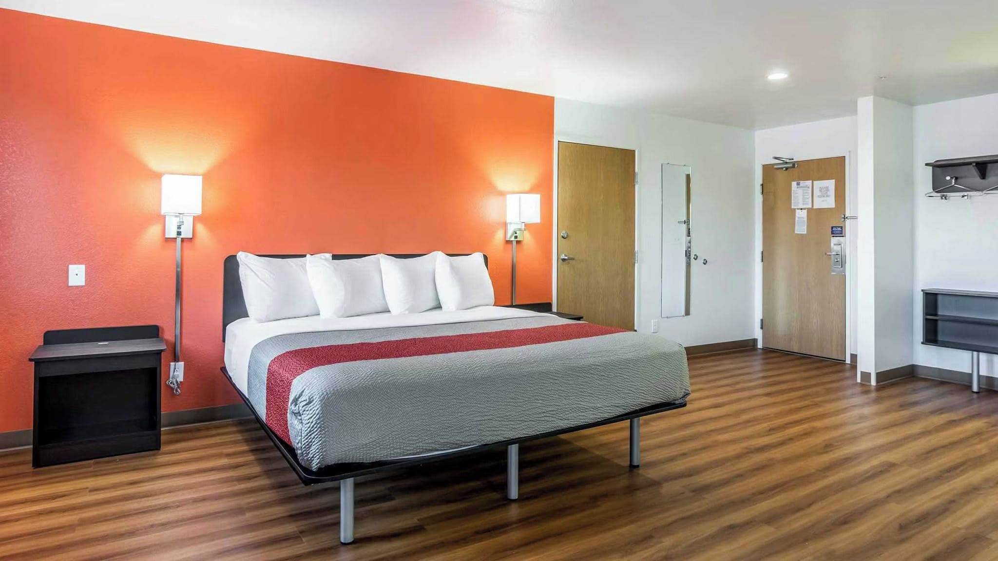 Una habitación de hotel con una cama, dos lámparas de noche, una puerta de madera, un perchero y un cubo de basura pequeño.