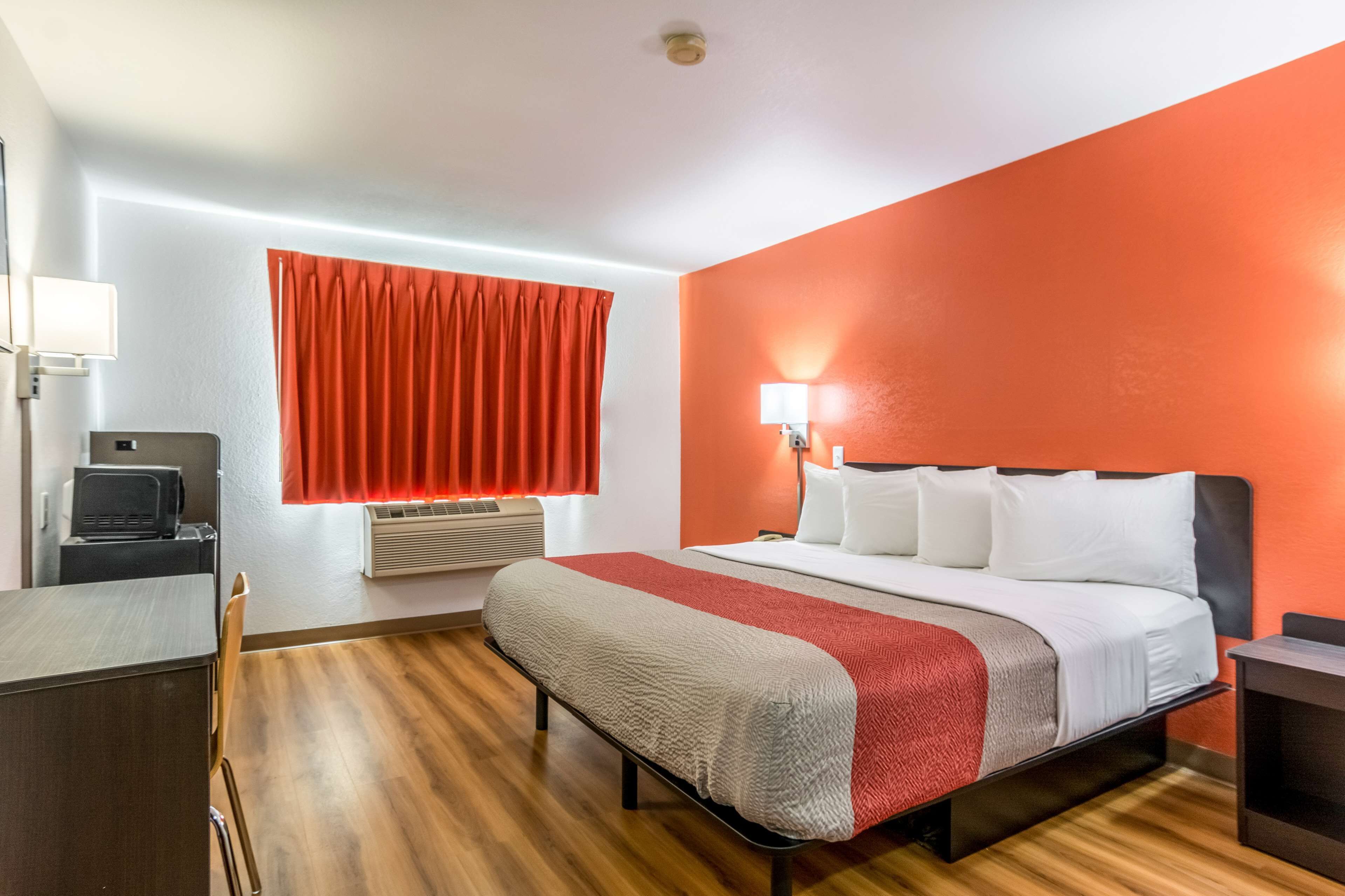 Habitación de hotel con cama, escritorio, silla, ventana con cortinas rojas y una lámpara de pared.