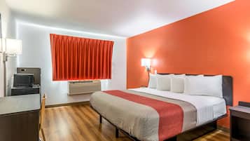 Habitación de hotel con cama, escritorio, silla, ventana con cortinas rojas y una lámpara de pared.