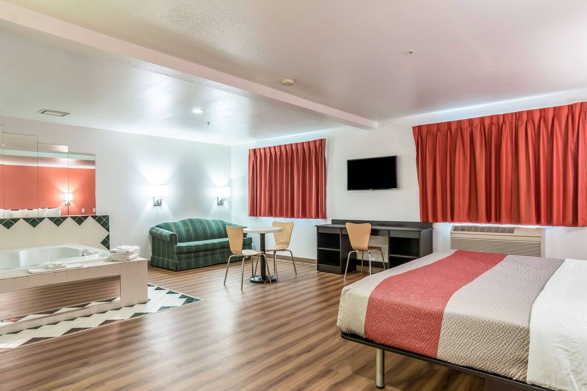 A hotel room with a bed, a desk, a chair, a sofa, a TV, a bathroom with a bathtub, and a window with curtains.