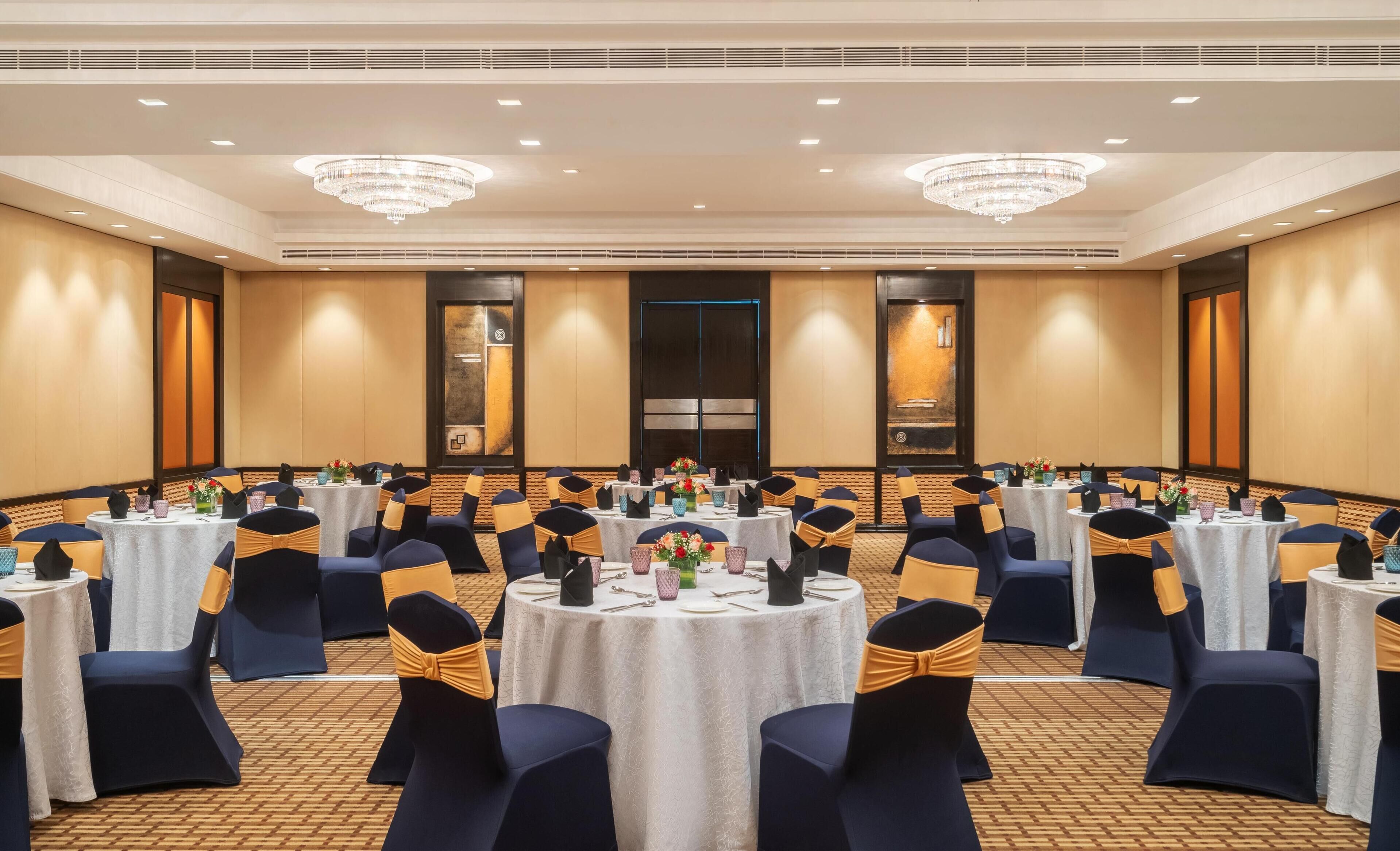 Un salón de conferencias con mesas redondas, sillas azul marino adornadas con cintas doradas y candelabros.