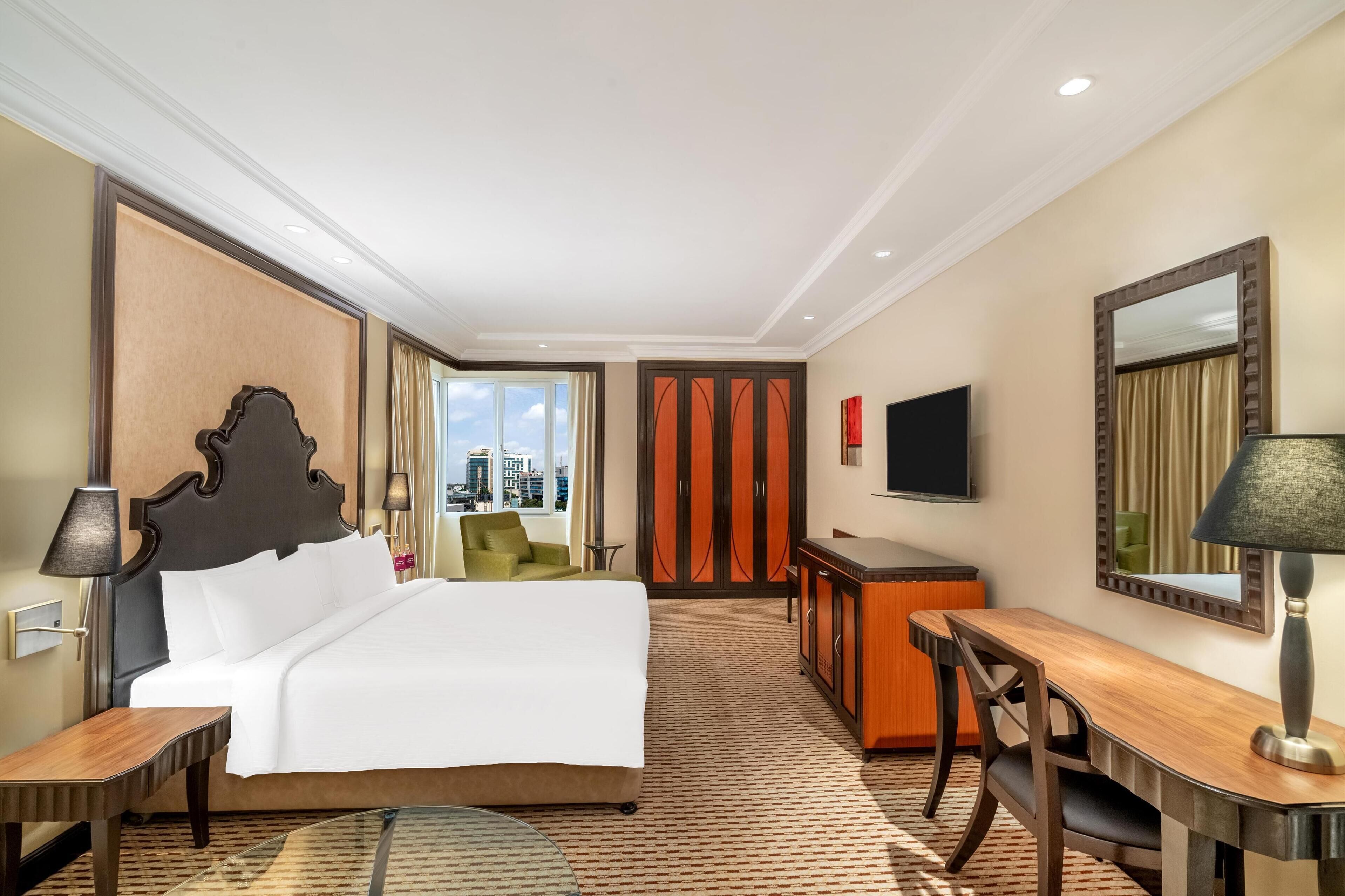 A hotel room with a large bed, a desk, a chair, a TV, and a window with a city view.