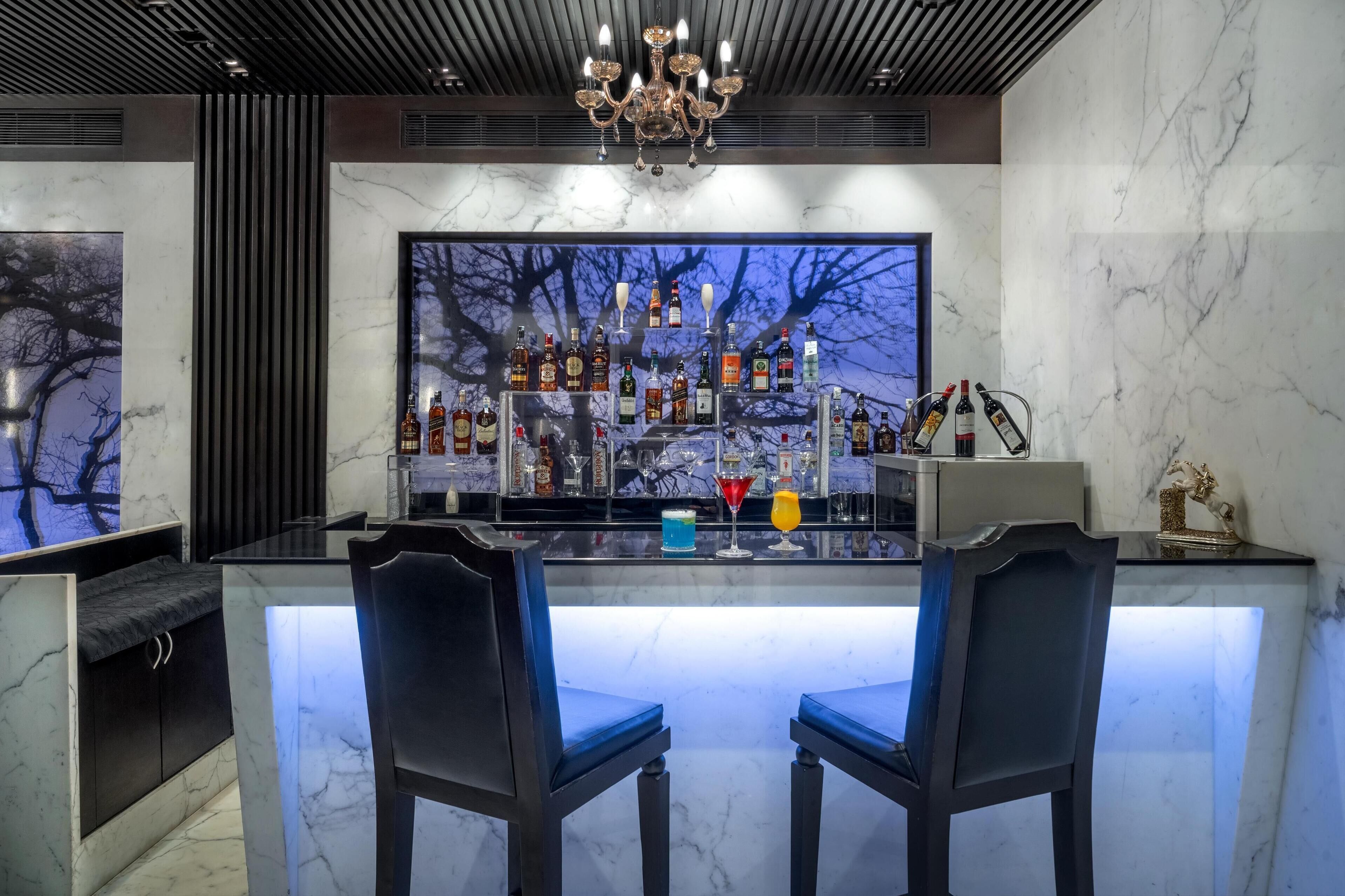 Een moderne bar met een marmeren aanrecht, twee barkrukken, een divers assortiment flessen en een groot raam met uitzicht op een boom.