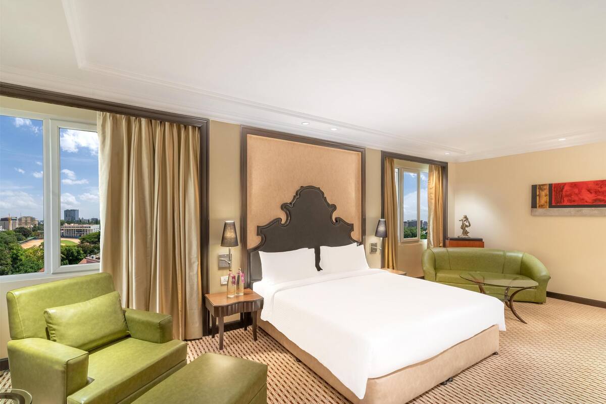 A hotel room with a large bed, a green armchair, a green sofa, a small table with a lamp, and a view of the city through large windows.