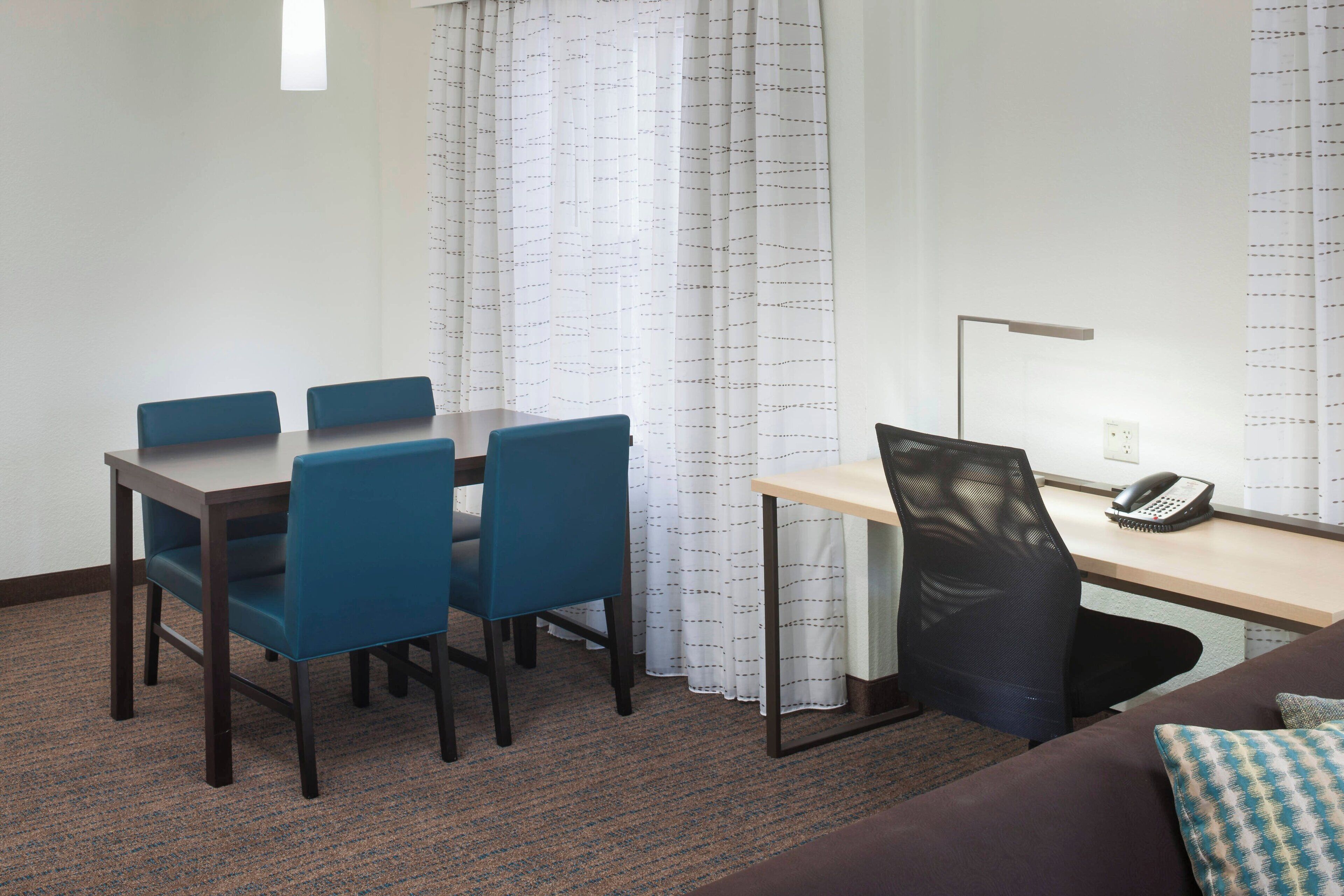 Habitación de hotel con una mesa de comedor, cuatro sillas azules, un escritorio con una silla, un teléfono y una ventana con cortinas.