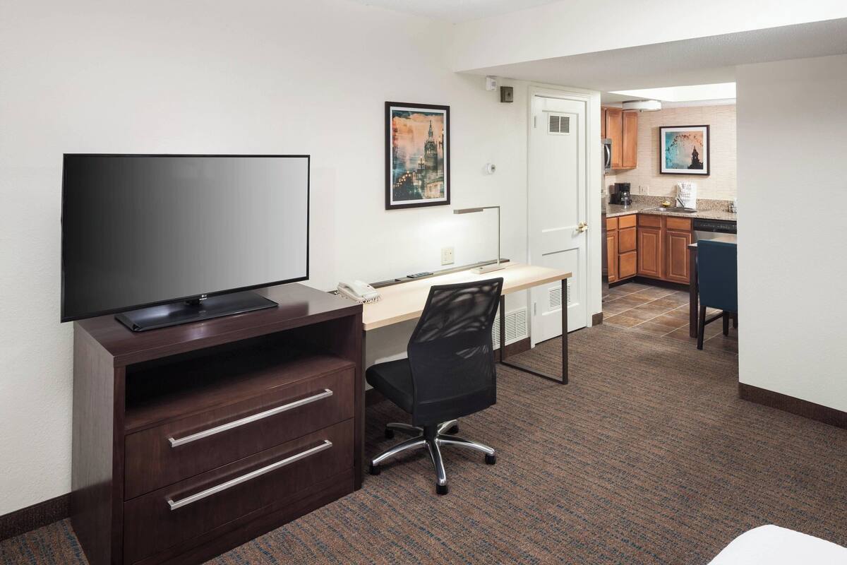 A hotel room with a flat-screen TV on a wooden cabinet, a desk with a chair, and a kitchenette in the background.