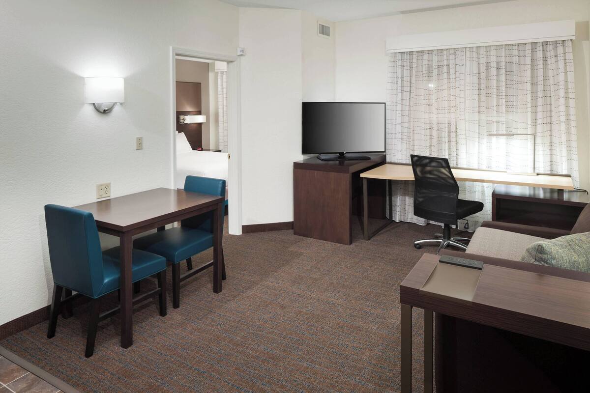 A compact hotel room with a small dining table, two blue chairs, a TV on a wooden stand, a desk with a chair, and a window with curtains.