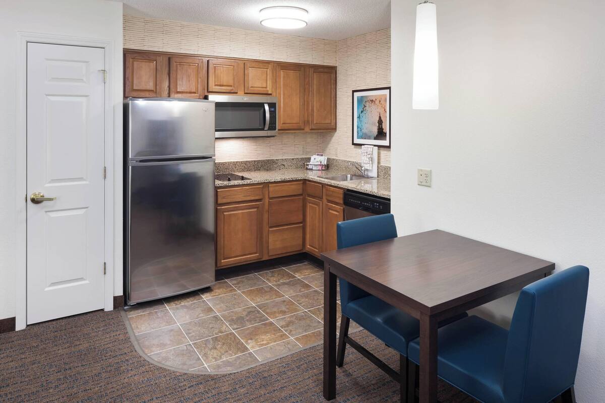 A compact kitchen with wooden cabinets, a stainless steel refrigerator, a microwave, and a dishwasher. There's a dining table with blue chairs.