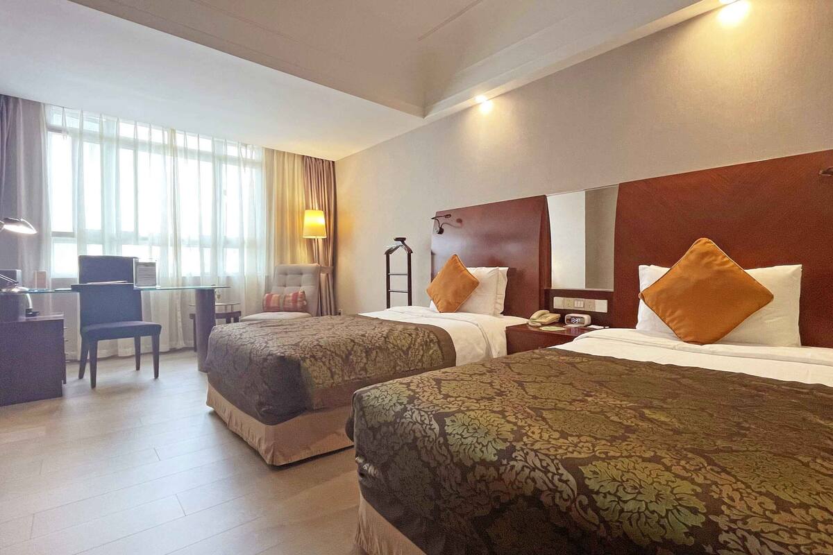 A hotel room with two beds, a desk, a chair, and a lamp.