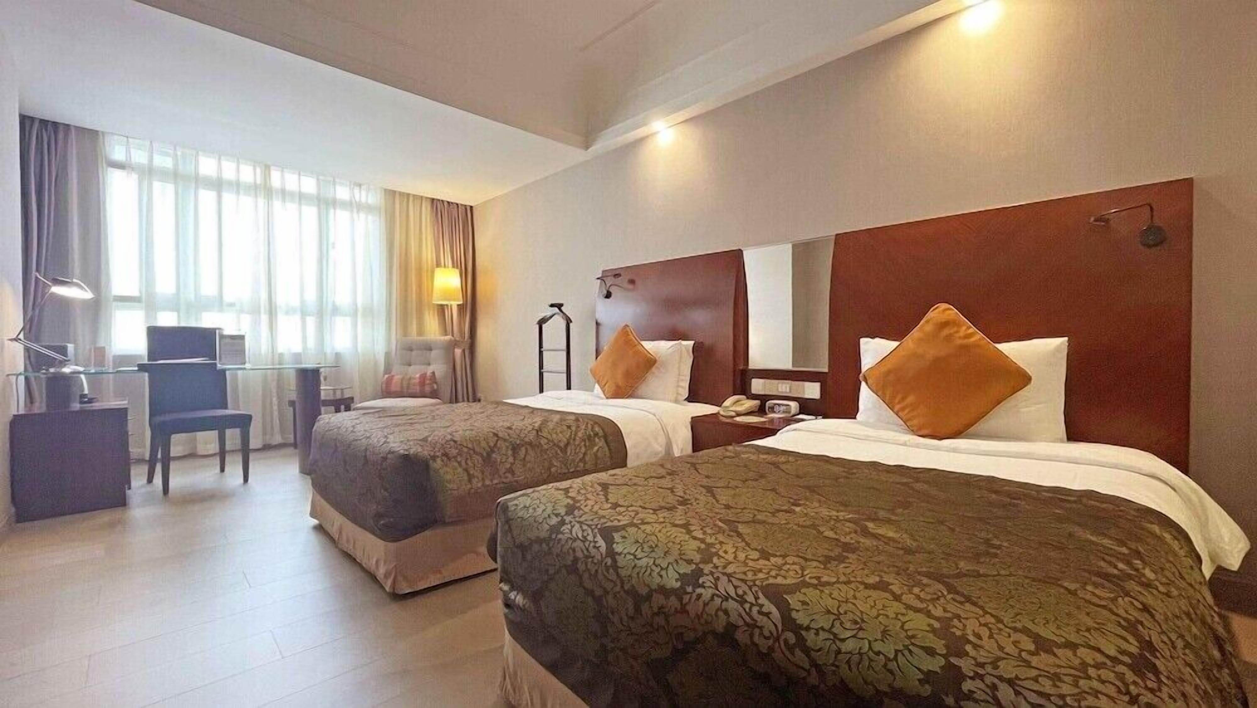 A hotel room with two beds, a desk, a chair, a lamp, and a window with curtains.