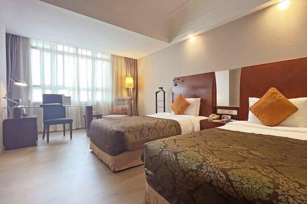A hotel room with two beds, a desk, a chair, a lamp, and a window with curtains.
