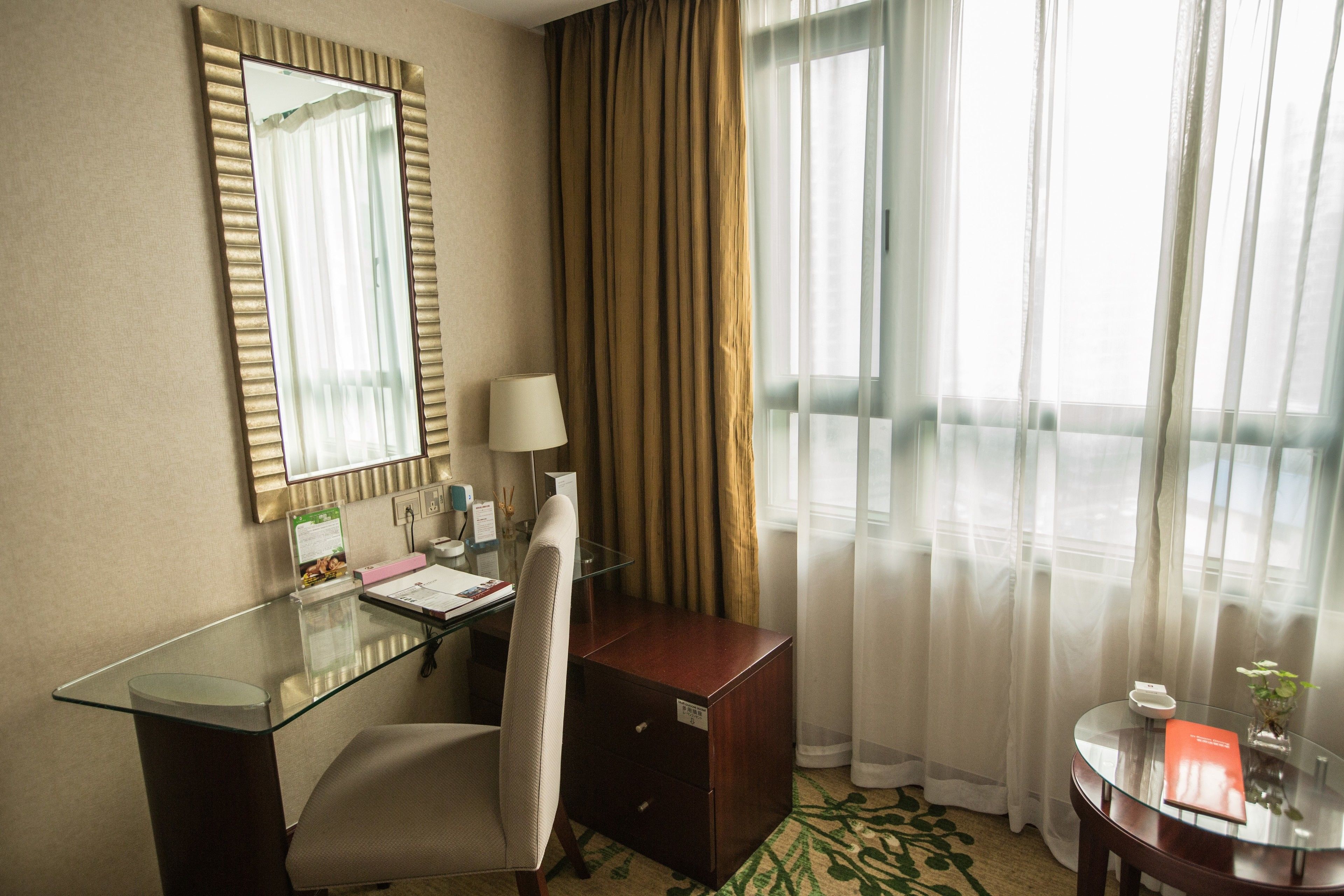 A hotel room with a glass desk, a chair, a wooden cabinet, a round table with a vase, and a large window with curtains.