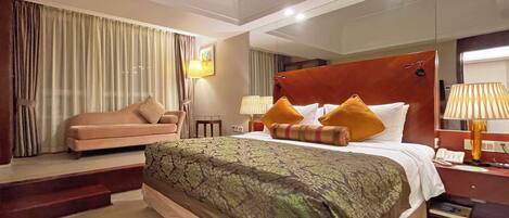 A hotel room with a large bed, a sofa, a desk with a lamp, and a mirror.