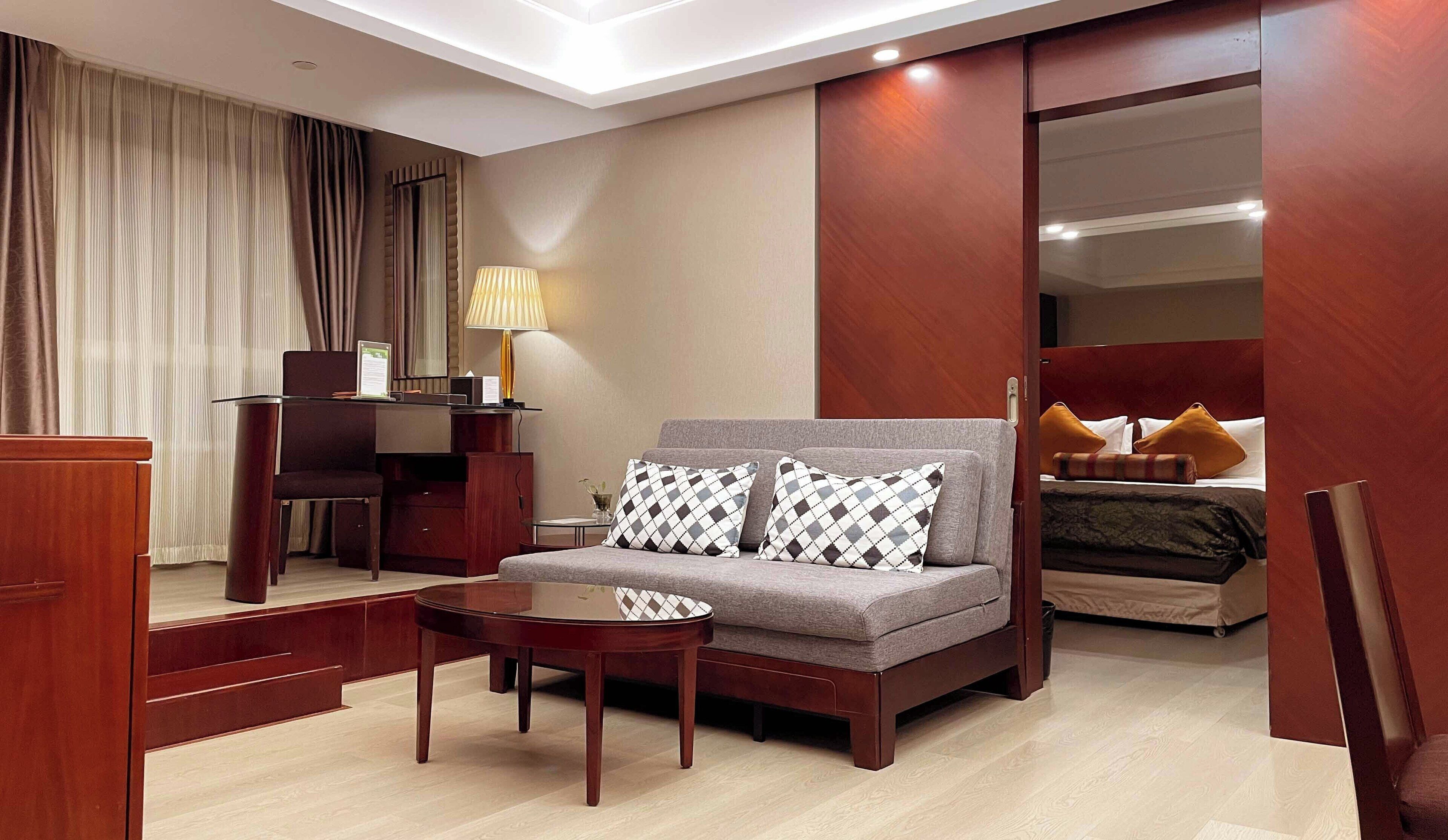 A modern hotel room with a sofa, a small round table, a desk with a lamp, and a bed with pillows.