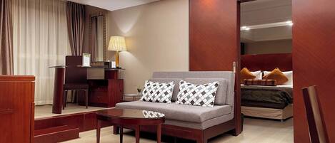 A modern hotel room with a sofa, a small round table, a desk with a lamp, and a bed with pillows.