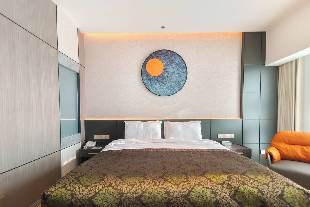 A modern hotel room with a large bed, a round artwork above the bed, a nightstand with a phone, and a seating area with a chair.