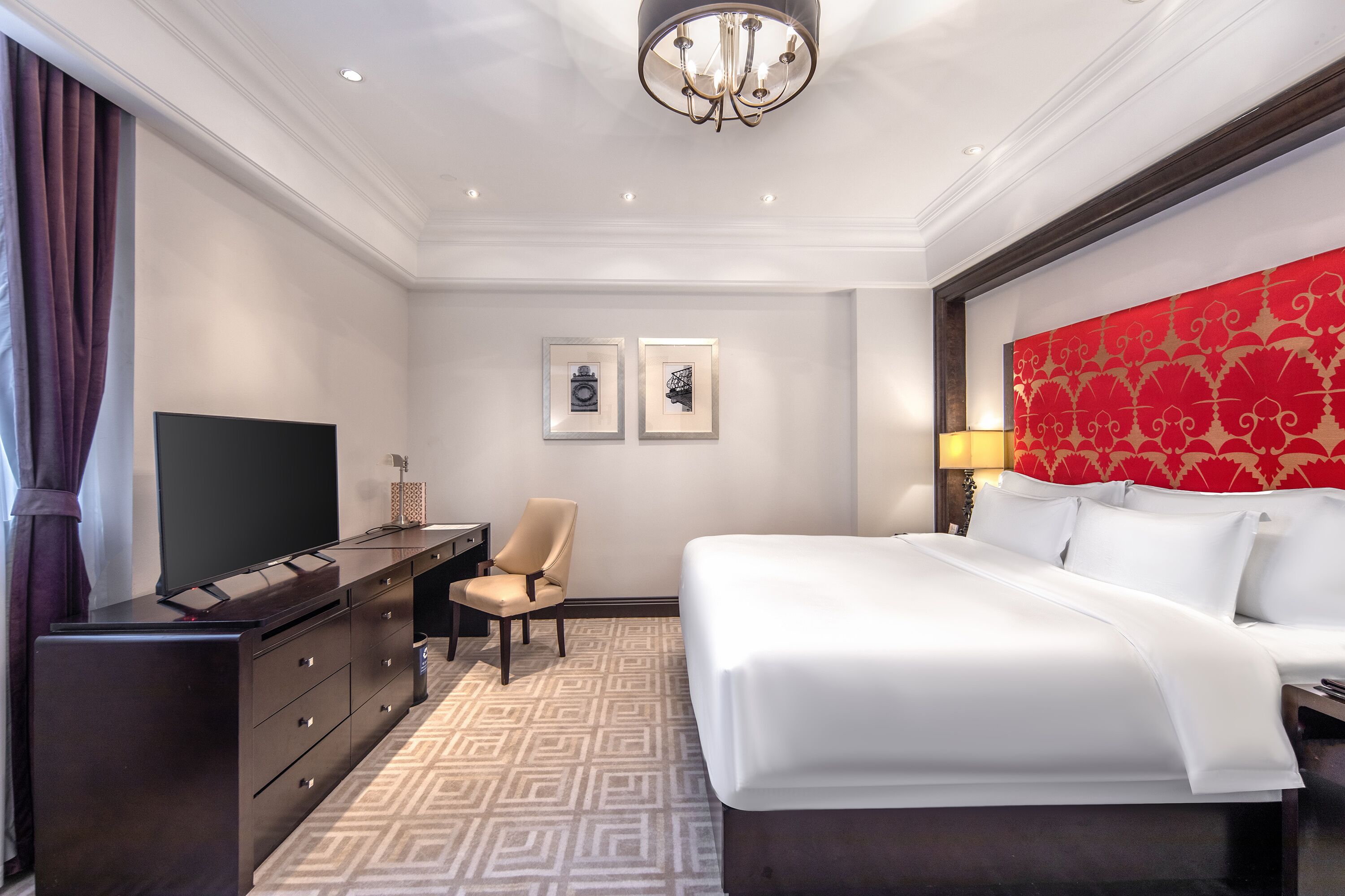 A hotel room with a large bed, a TV, a desk, and a chair. There are two framed pictures on the wall.