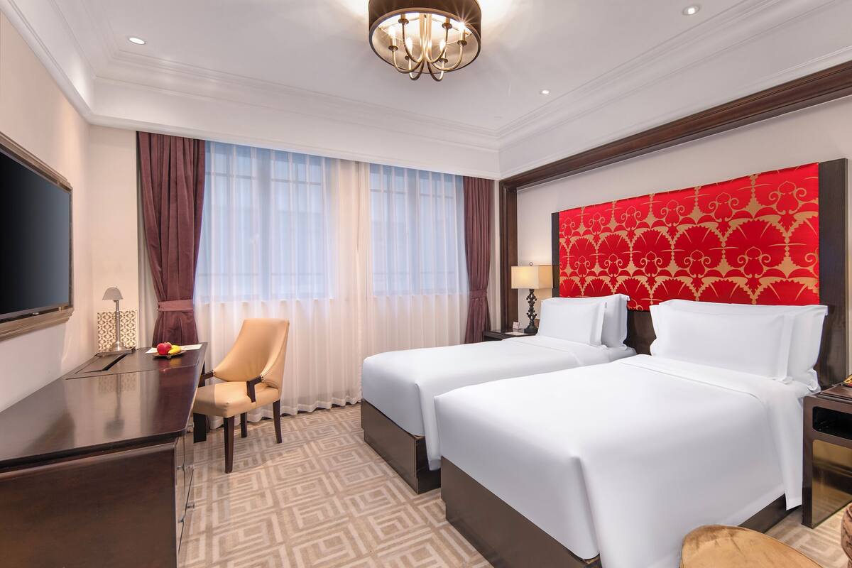A hotel room with two beds, a desk, a chair, a TV, and a chandelier.