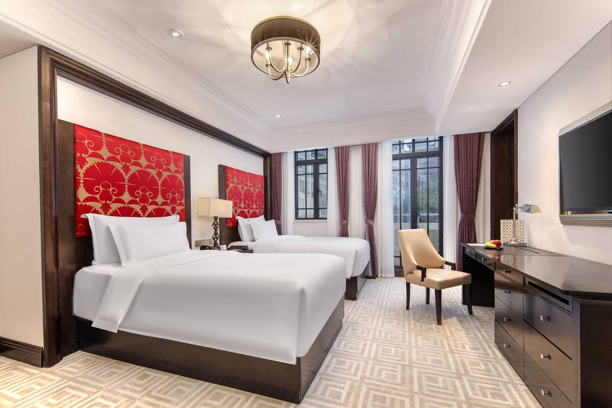 A hotel room with two beds, a desk, a chair, a TV, and large windows with curtains.