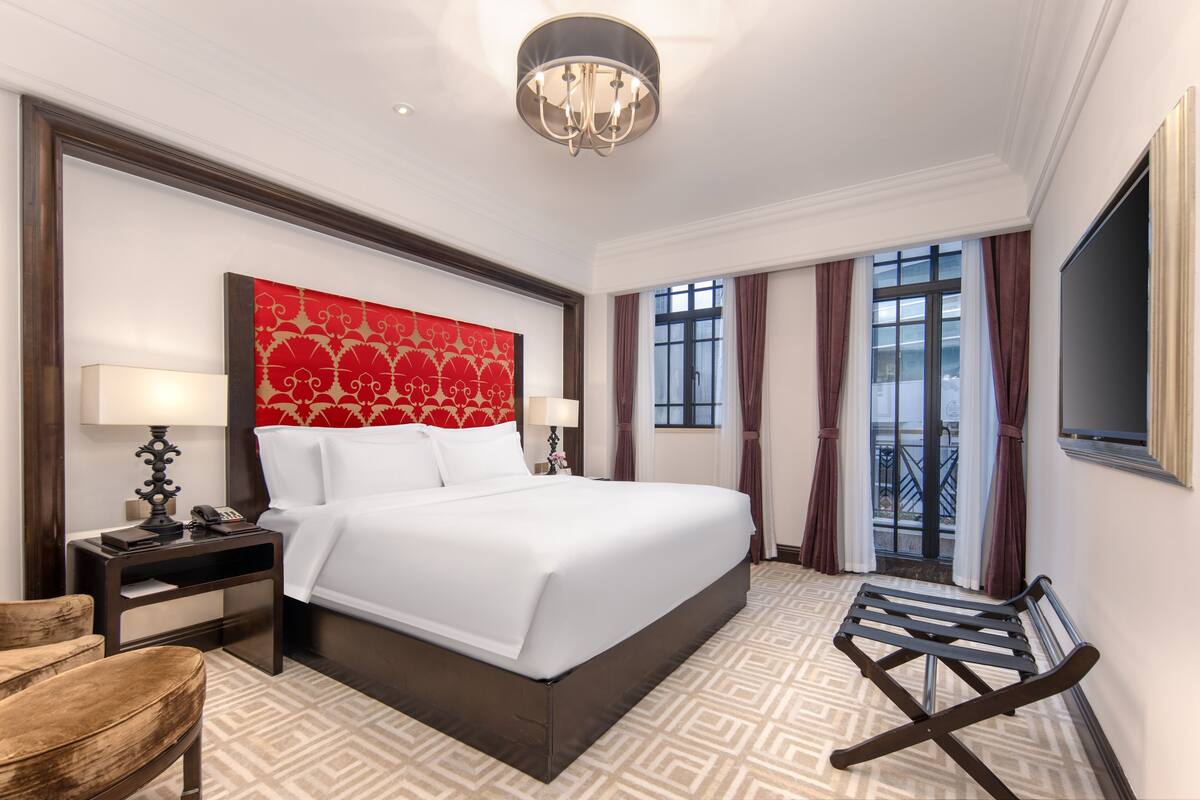 A hotel room with a large bed, two bedside tables with lamps, a red patterned headboard, a large window with curtains, and a TV mounted on the wall.