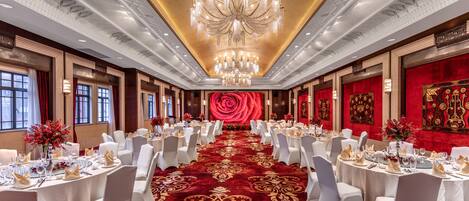 Une salle de banquet avec des tables rondes, des chaises blanches, une moquette rouge et un lustre.
