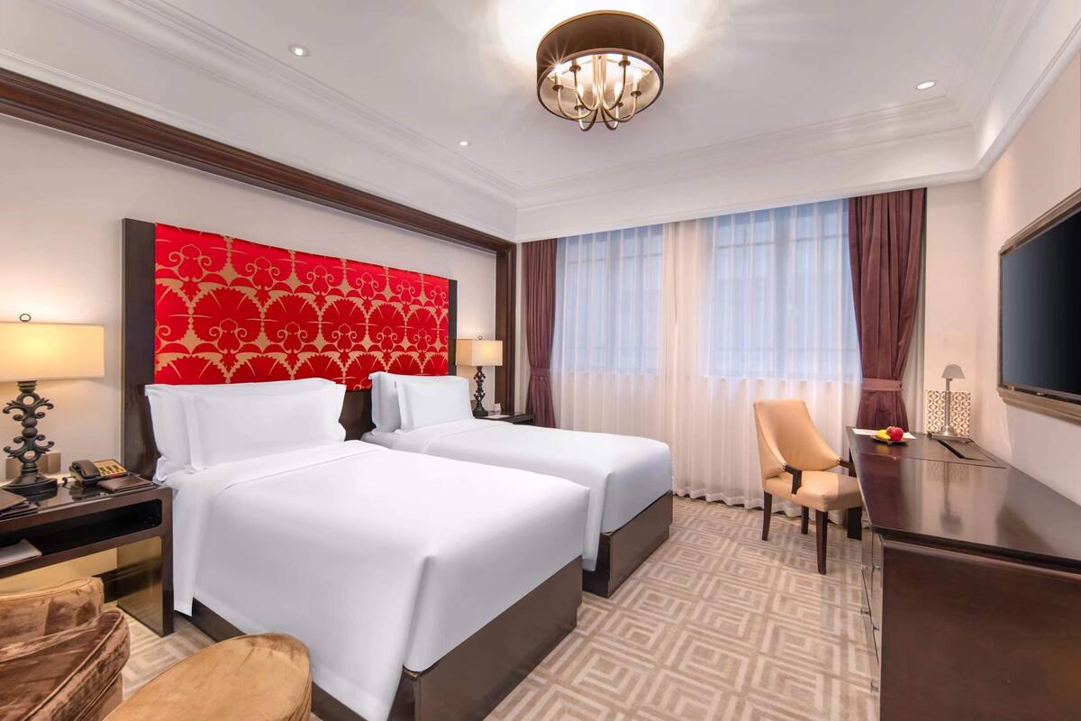 A hotel room with two beds, a desk, a chair, a TV, and a large red patterned wall.