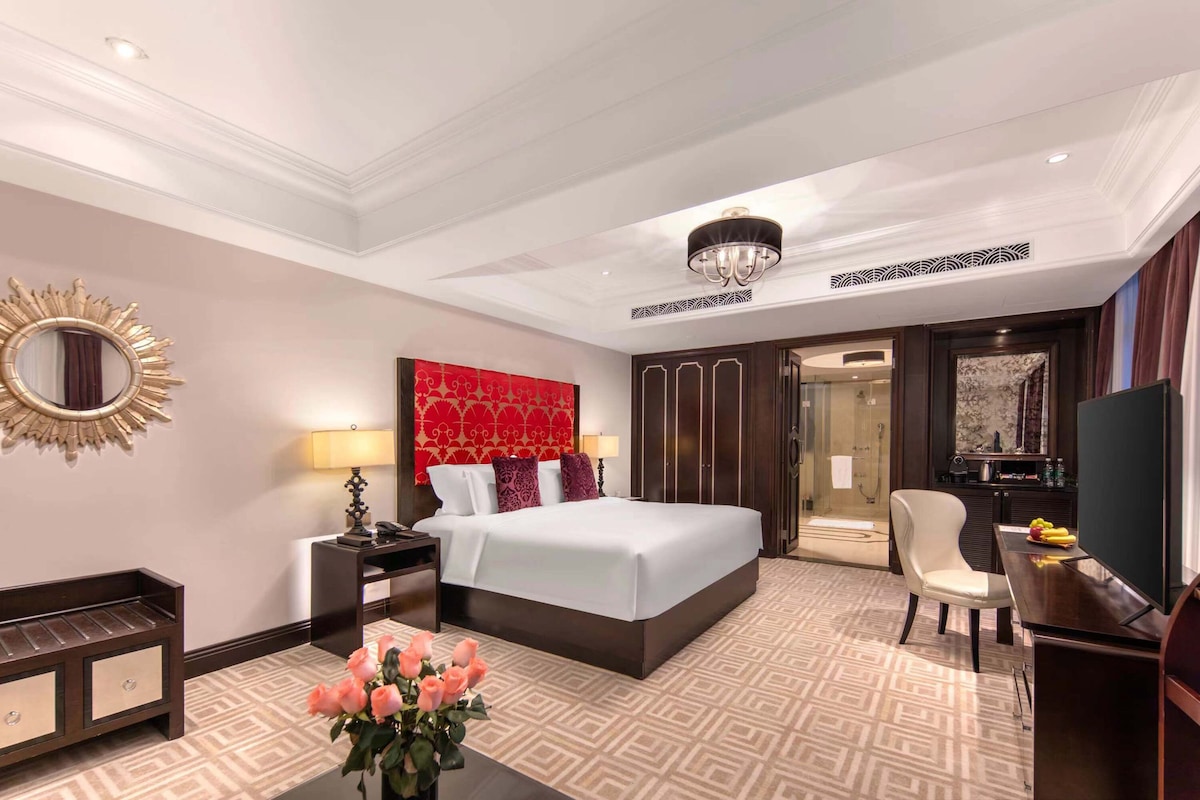 A hotel room with a large bed, a sitting area with a chair and a table, a vanity with a mirror, and a television.