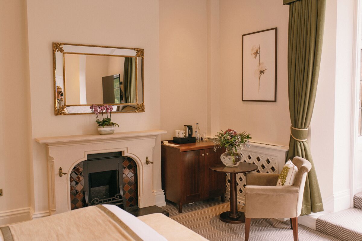 A hotel room with a fireplace, a mirror, a desk with a vase of flowers, a chair, and a framed picture on the wall.