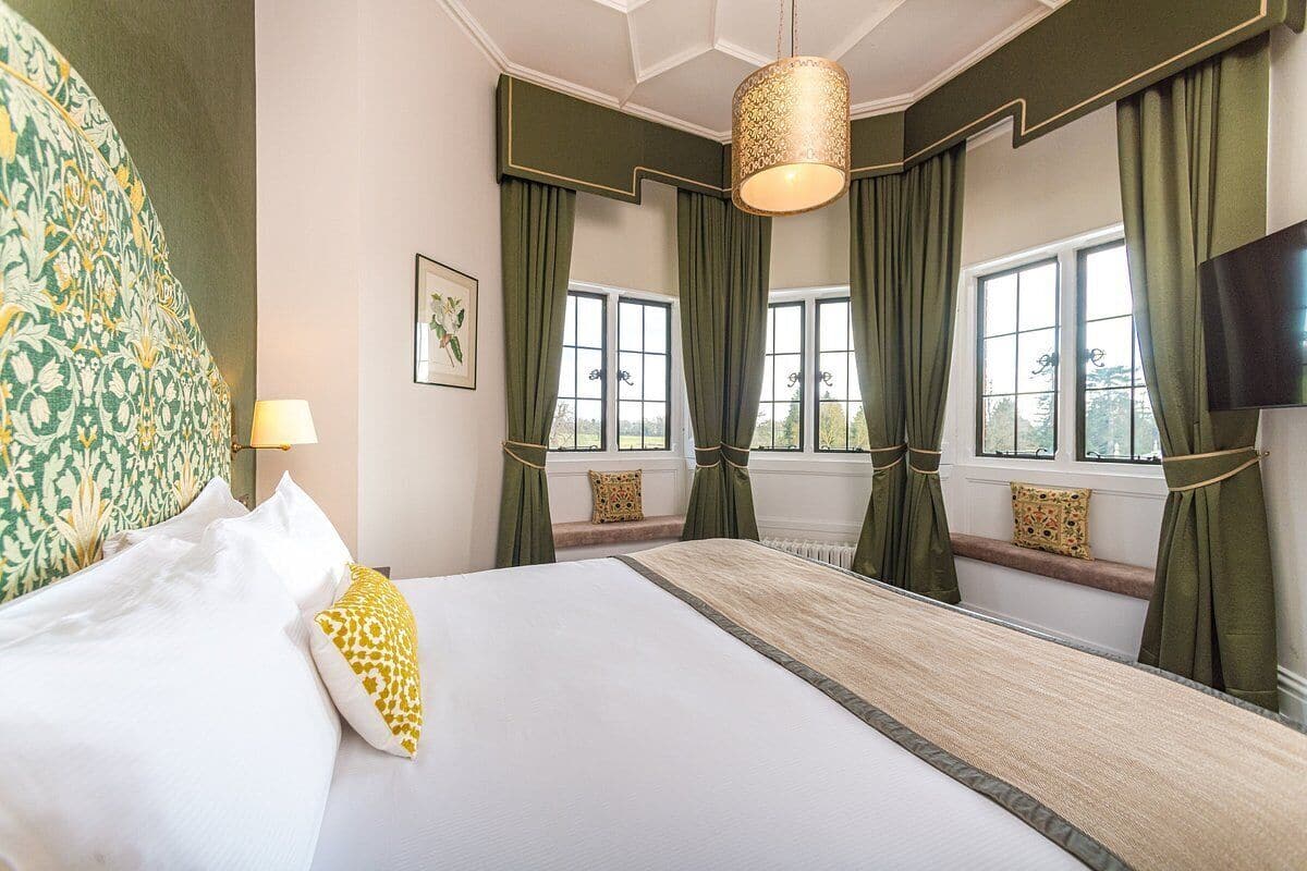 Une chambre d’hôtel avec un grand lit, des rideaux verts, une lampe suspendue et une vue sur l’extérieur par les fenêtres.