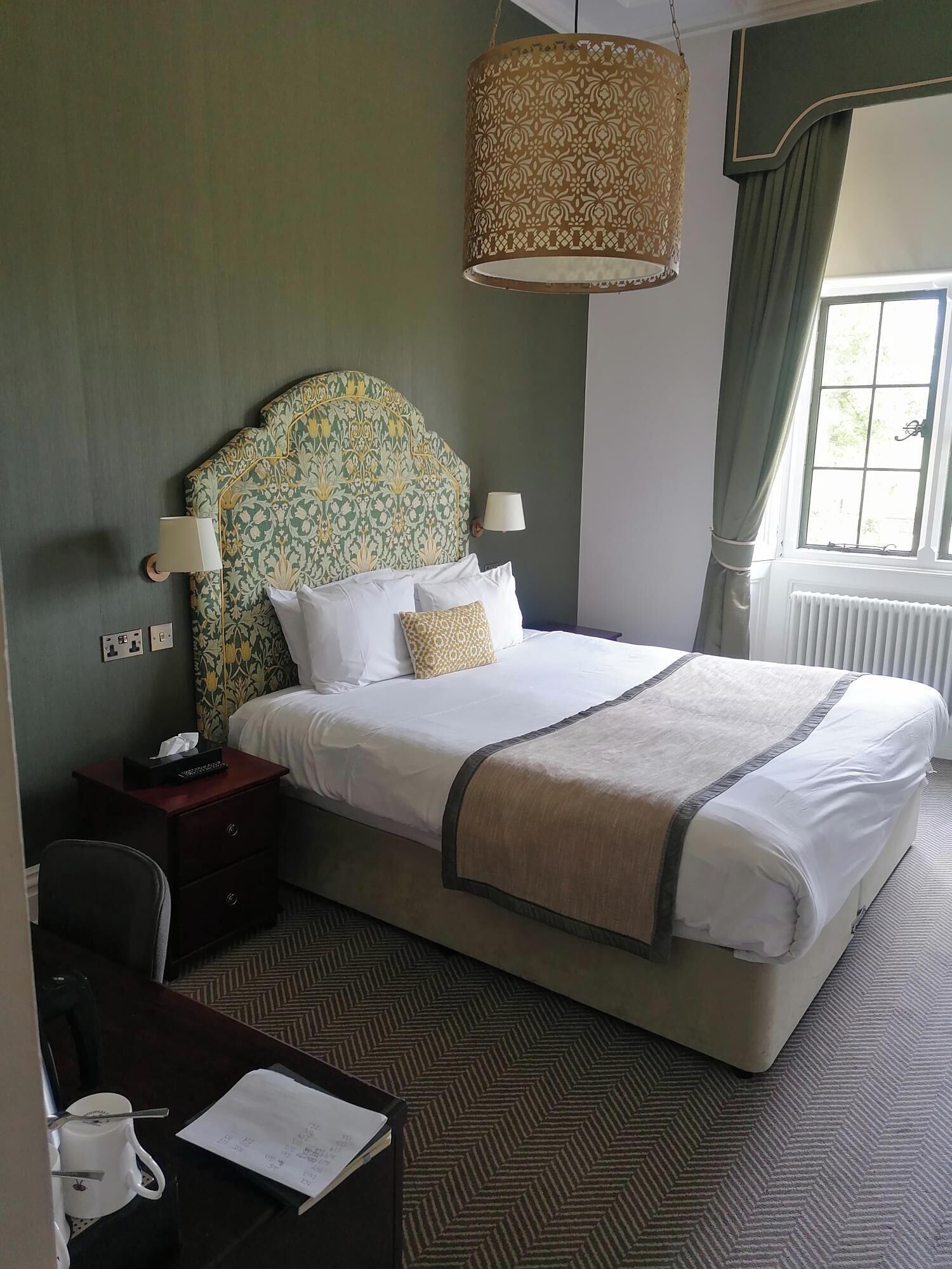 A hotel room with a large bed, two bedside lamps, a patterned headboard, a hanging lamp, a window with curtains, and a desk with a cup and papers.