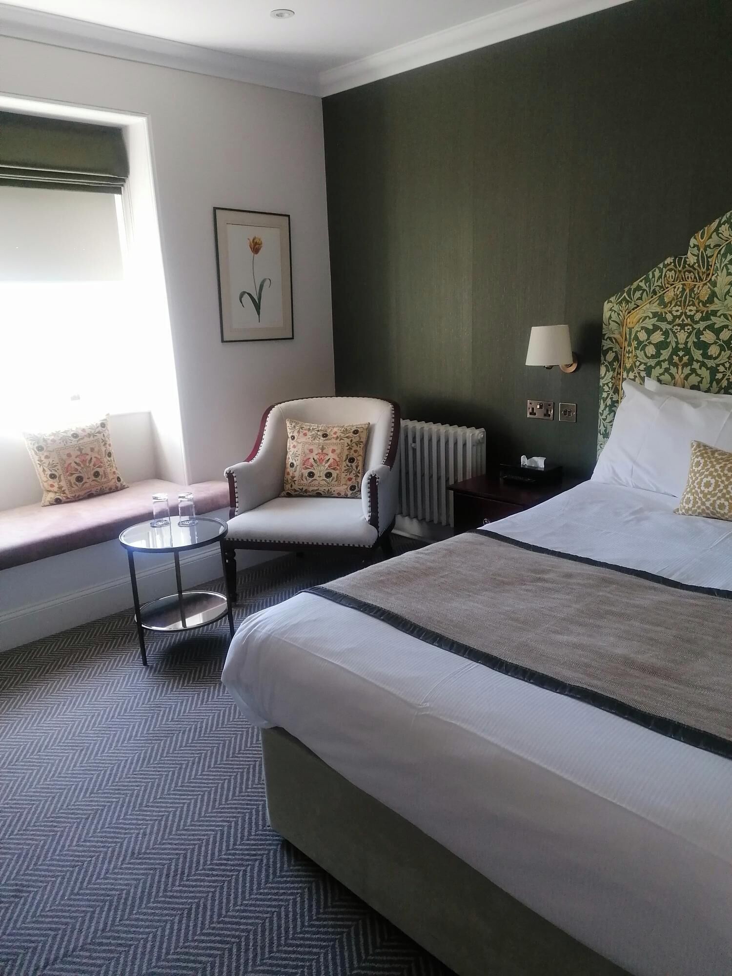 A hotel room with a bed, a chair, a small table, a radiator, and a window with a view.