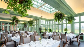Un amplio comedor interior con varias mesas dispuestas para comer, rodeado de grandes ventanales y plantas verdes.