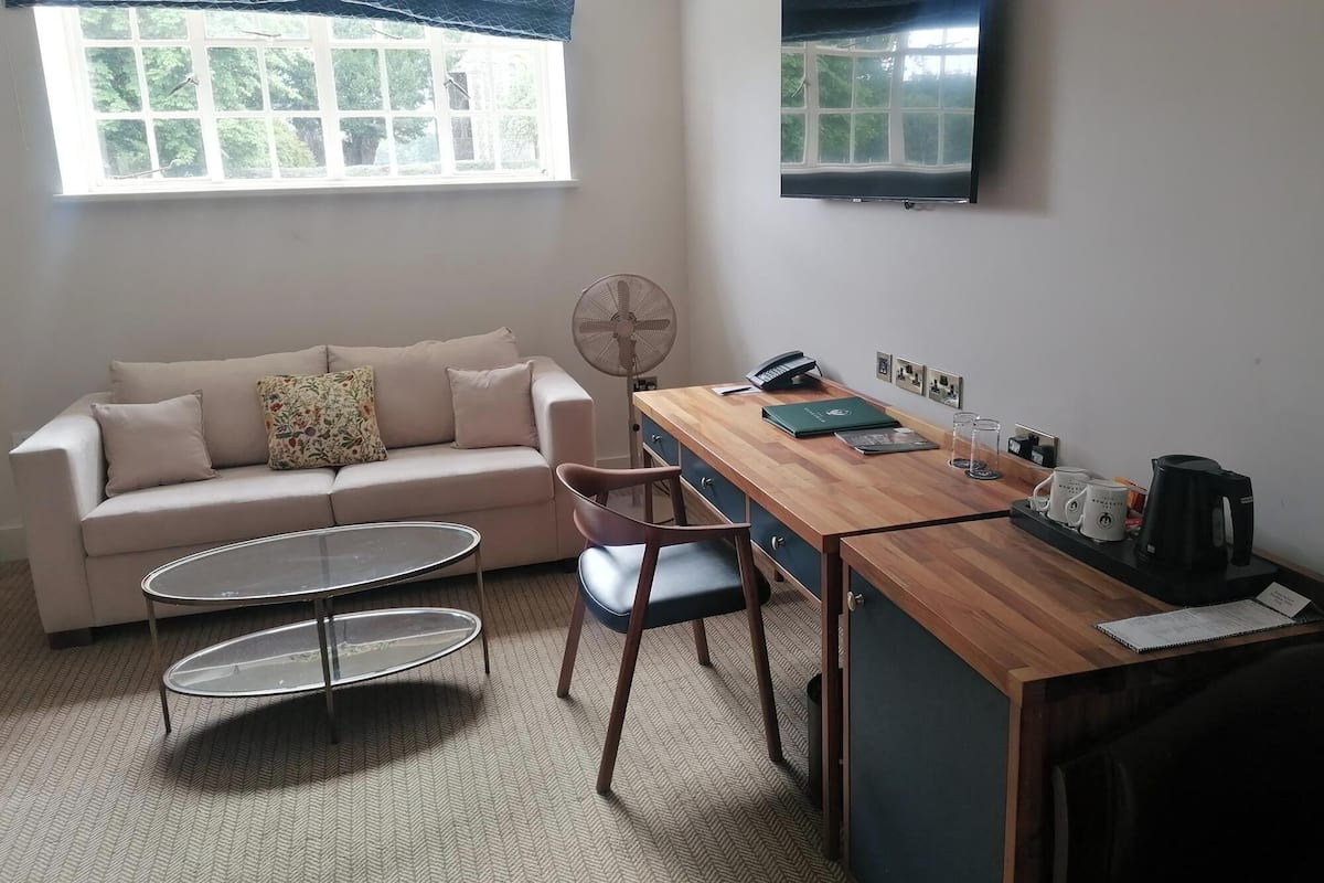 Une petite pièce bien éclairée, avec un canapé, une table basse, un bureau avec une chaise, un ventilateur, un téléviseur et une fenêtre donnant sur des arbres.