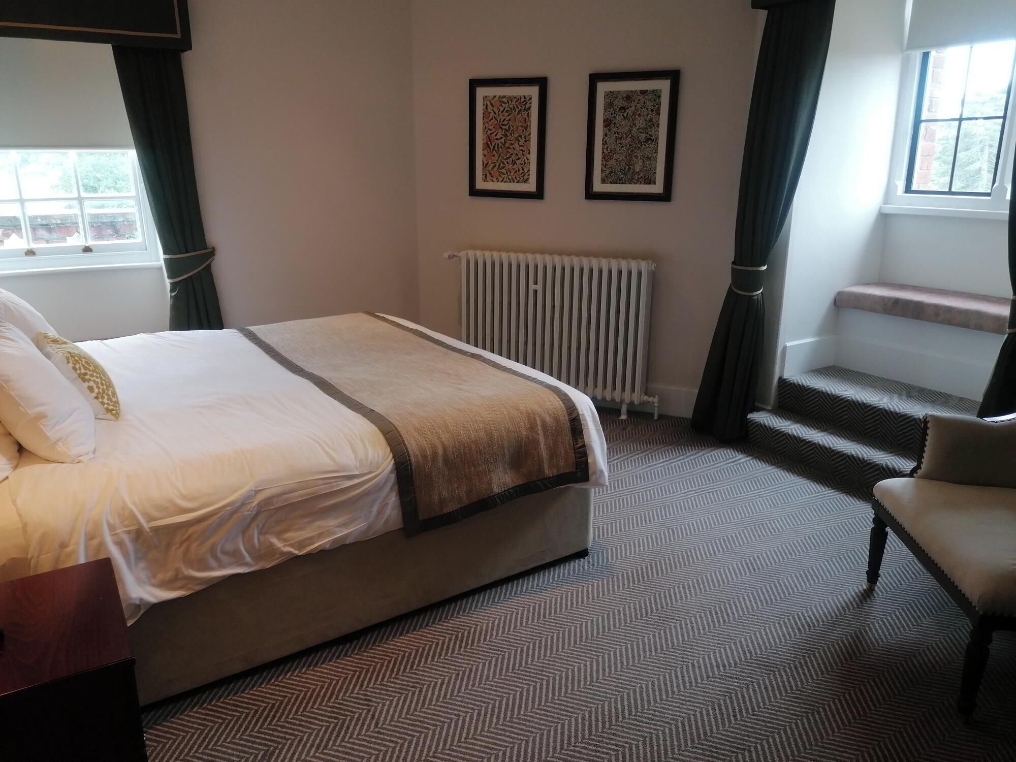 A hotel room with a bed, a chair, a radiator, and two framed pictures on the wall.