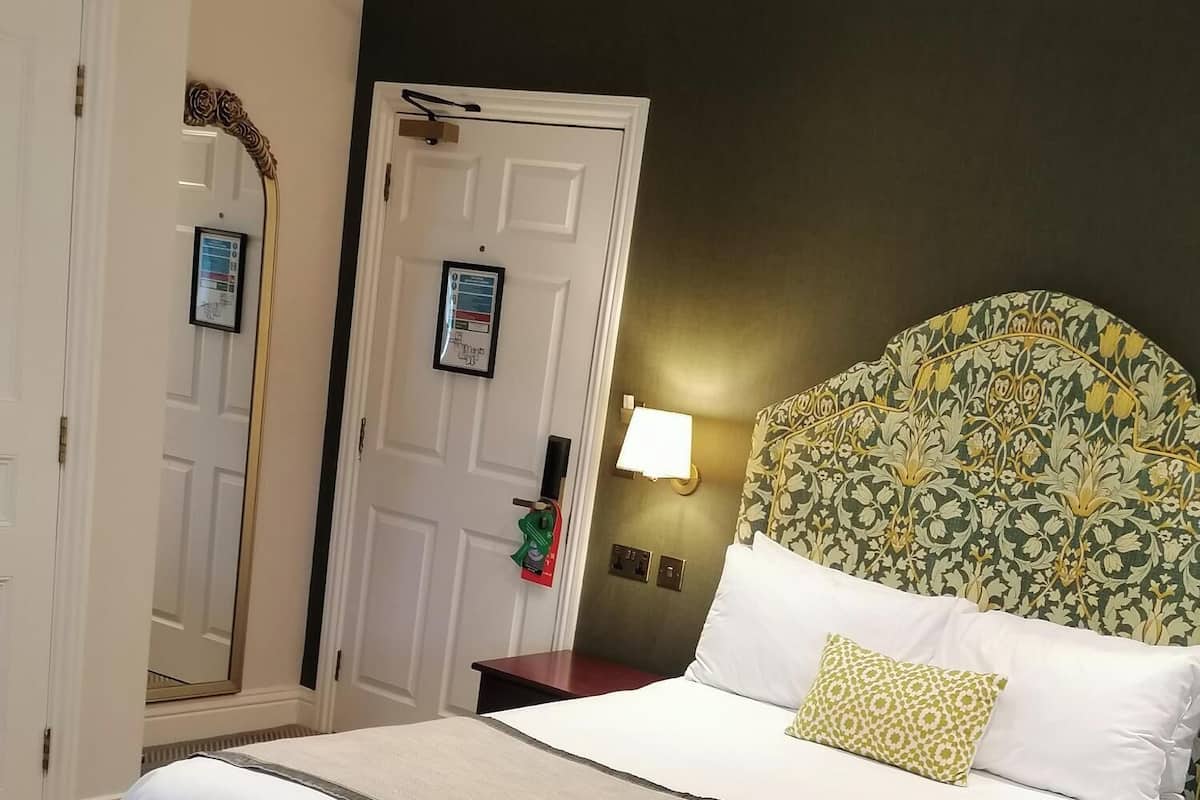 Une chambre d’hôtel avec un grand lit, une tête de lit ornée d’un motif floral, une table de chevet avec une lampe et une porte dotée d’un miroir et d’un détecteur d’incendie.