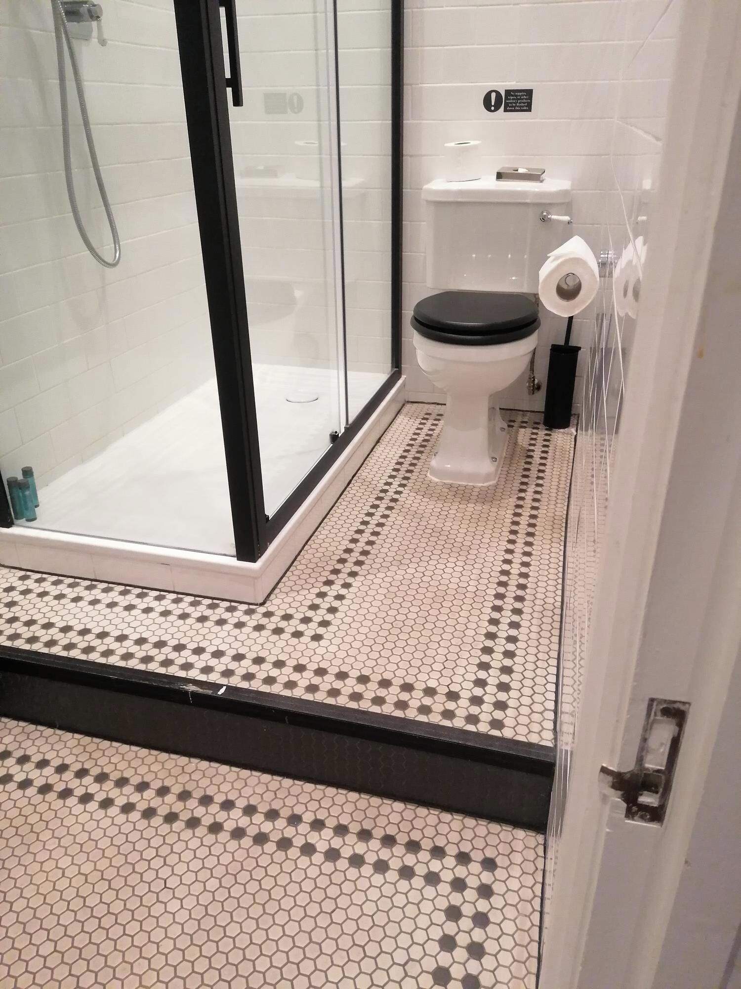 Un baño con cabina de ducha de vidrio, inodoro con asiento negro y suelo de baldosas con patrón hexagonal.