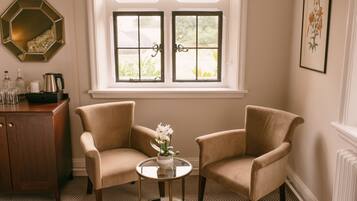 Una habitación con dos sillones beige, una mesita redonda con un florero, un armario de madera con botellas y una ventana que da a zonas verdes.
